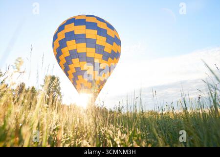 Montgolfière s'élevant au-dessus d'un champ herbeux avec la lumière du soleil qui brille à travers, motif à carreaux bleu et jaune visible, arbres et ciel en arrière-plan, concept d'aventure en plein air Banque D'Images