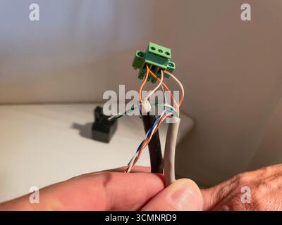 Gros plan d'une main tenant un bornier vert avec trois fils (orange, blanc et vert) connectés, émergeant d'un câble gris, contre une lumière b. Banque D'Images