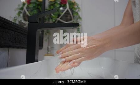 Femme se lavant les mains sous l'eau courante à un robinet noir élégant dans une salle de bain lumineuse. Banque D'Images