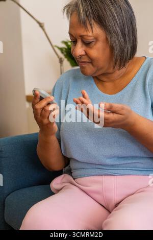 Femme afro-américaine senior tenant un glucomètre et vérifiant la main tout en étant assise dans le salon Banque D'Images