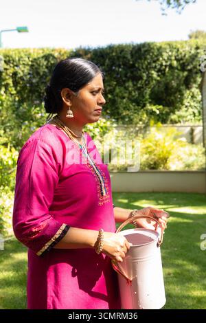 Femme indienne portant kurta rose et arrosage de plantes avec arrosoir dans la cour ensoleillée, espace de copie Banque D'Images