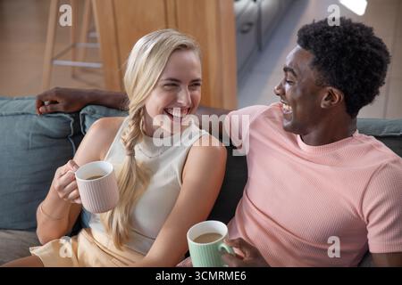 Couple diversifié assis sur le canapé sirotant le café des tasses et riant dans le salon moderne Banque D'Images
