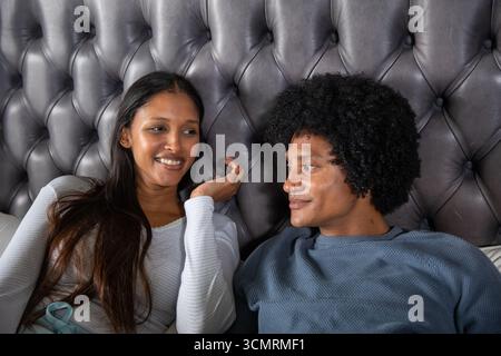 Couple diversifié parlant tout en étant allongé sur le lit dans la chambre, tête de lit grise touffetée, oreillers Banque D'Images