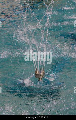 Vue détaillée d'une buse de fontaine pulvérisant plusieurs jets d'eau dans une piscine turquoise, créant des éclaboussures et des ondulations sous la lumière du jour. Banque D'Images