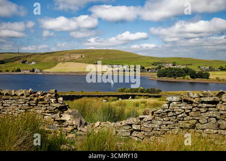 Un espace dans un mur de pierre sèche encadre une vue du réservoir de Clowbridge avec un seul yacht à voile shite et des moutons se reposant à l'ombre d'un mur, Burnley. Banque D'Images