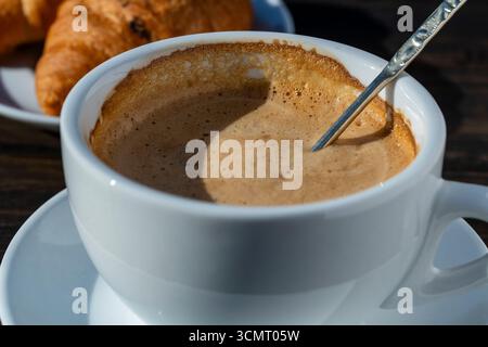 Mousse épaisse de cappuccino de café chaud avec cuillère dans une tasse blanche et croissant sur une table en bois, gros plan. Cappuccino mousse de lait, latte Banque D'Images