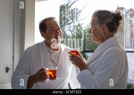 Couple de personnes âgées debout tenant des tasses de thé en verre dans des peignoirs au spa près du mur de verre Banque D'Images