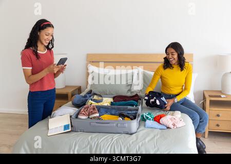 Maman diversifiée et fille adolescente tenant le téléphone et pliant les vêtements de la valise sur le lit Banque D'Images