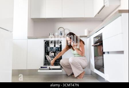 Jeune femme à la maison cuisine appelant le service parce que la machine à laver est cassée Banque D'Images