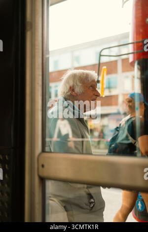 Twickenham, Londres, Royaume-Uni – 2025.09.06 – un homme âgé aux cheveux blancs attend devant un bus rouge londonien, vu à travers une fenêtre d’arrêt de bus avec des reflets de passe Banque D'Images