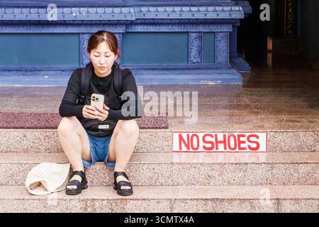 Jeune femme asiatique saisissant des chaussures assis sur les marches du temple en utilisant un smartphone à côté de pas de chaussures signe batu grottes, kuala lumpur malaisie Banque D'Images