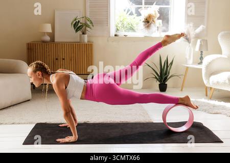 Femme effectue une planche avancée en utilisant une roue de yoga pour construire l'équilibre de force de base et la mobilité dans un studio à domicile lumineux Banque D'Images