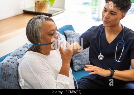 Femme afro-américaine senior utilisant un masque à oxygène, infirmière asiatique en gommage guidant Banque D'Images