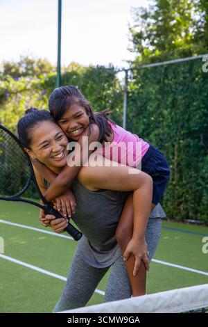 Mère asiatique et fille en vêtements de sport donnant piggyback ride sur court de tennis tout en tenant la raquette Banque D'Images
