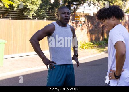 Divers amis masculins s'arrêtant et discutant à côté de la clôture, poubelle verte sur la rue de banlieue Banque D'Images