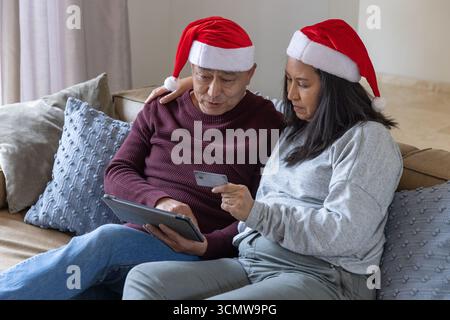 Couple senior diversifié dans chapeaux de père noël assis sur le canapé à la maison navigation tablette avec carte de crédit Banque D'Images