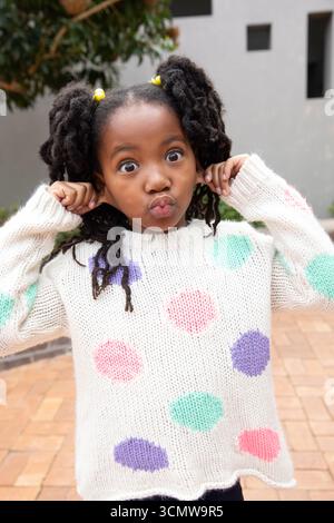Fille afro-américaine tirant les oreilles et plongeant les lèvres sur la cour en briques dans un pull à pois Banque D'Images