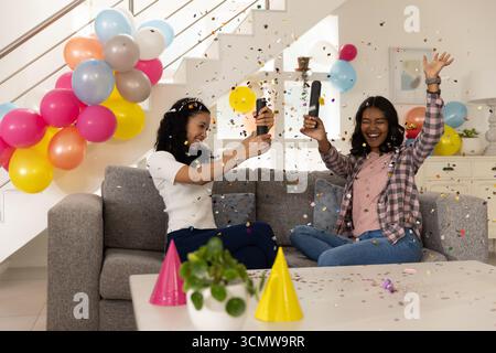 Maman diversifiée et fille adolescente éclatant des canons à confettis sur un canapé gris dans le salon Banque D'Images