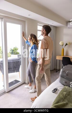 Couple debout par la porte coulissante en verre donnant sur le patio ensemble ; valise avec lunettes de soleil sur le lit Banque D'Images