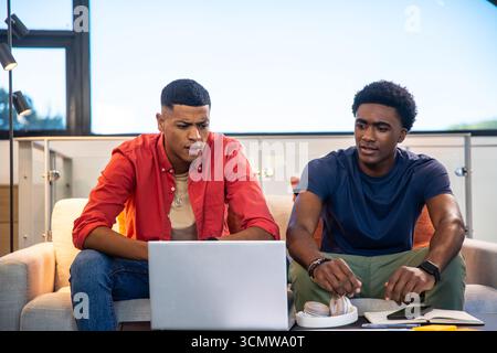 Divers amis masculins assis sur le canapé et collaborant sur ordinateur portable dans le salon avec des écouteurs Banque D'Images