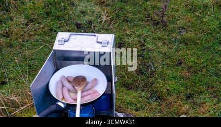 Saucisses frit dans une poêle sur un poêle de camping portable à l'extérieur Banque D'Images
