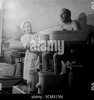 Une photographie historique des années 1960-1970, prise dans un sanatorium de Slavkurort, en RSS d'Ukraine, montrant un patient masculin subissant une procédure de physiothérapie - un bain galvanique à quatre chambres. Une infirmière supervise le processus de traitement dans le balnéaire, illustrant l'approche scientifique de la réadaptation et de l'amélioration de la santé des travailleurs au sein du système de santé soviétique. La photo capture l'atmosphère de la pratique médicale et des soins aux patients dans la célèbre station de santé de Donbass Banque D'Images
