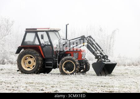 Un tracteur rouge est stationnaire dans un champ couvert de gel entouré d'arbres enneigés et de brume. Banque D'Images