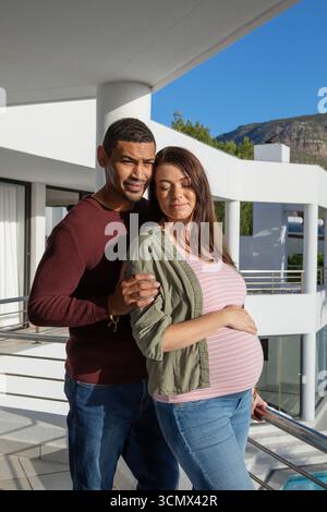 Partenaires féminins divers debout sur le balcon moderne tenant le ventre enceinte près de la balustrade métallique Banque D'Images