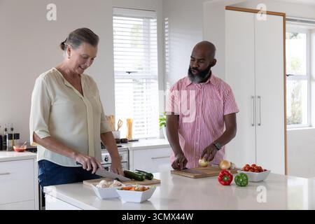 Couple senior diversifié hachant des légumes sur l'îlot de cuisine avec des planches à découper en bois et des couteaux Banque D'Images