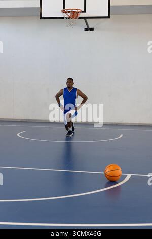 Homme afro-américain s'étirant en fente avant sur le terrain bleu portant un maillot avec balle et cerceau Banque D'Images