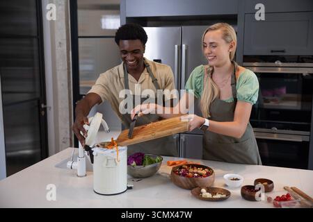 Couple diversifié hachant des légumes sur une planche à découper dans la cuisine à la maison avec des bols, bac à compost Banque D'Images