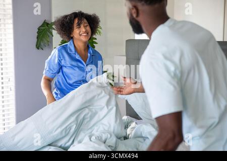 Couple diversifié en pyjama redressant housse de couette bleu pâle sur le lit dans la chambre Banque D'Images