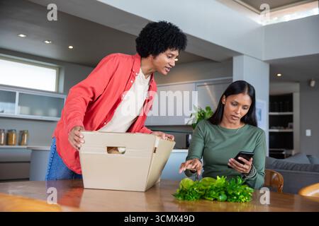 Couple diversifié déballer la boîte en carton et examiner les verts feuillus, en utilisant un smartphone dans la cuisine Banque D'Images
