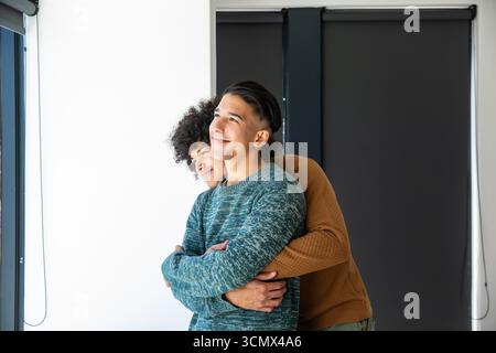 Divers partenaires masculins embrassant et regardant à travers une grande fenêtre avec abat-jour rouleau dans le salon Banque D'Images