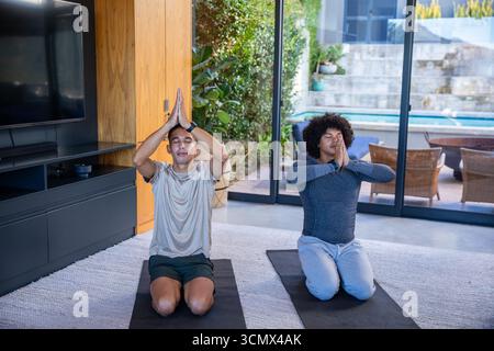 Divers amis masculins agenouillant sur des tapis de yoga et méditant dans le salon à côté de la porte coulissante en verre Banque D'Images