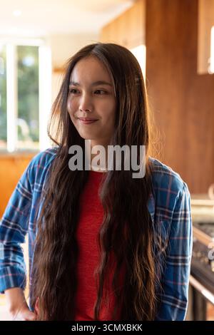 Femme asiatique portant une chemise à carreaux, debout dans la cuisine, regardant vers la fenêtre par les armoires Banque D'Images