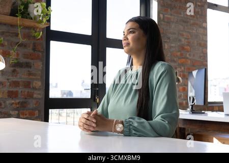 Femme assise au bureau blanc dans le bureau de style loft portant chemisier vert sauge, vérifier l'ordinateur portable Banque D'Images