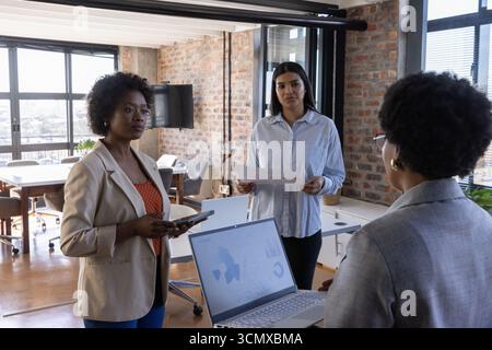Diverses collègues féminines examinant les ordinateurs portables, tablettes et documents imprimés dans un bureau moderne ouvert Banque D'Images
