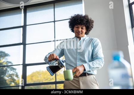 Homme versant le café du pot en verre dans la tasse verte par la fenêtre de grille dans la zone de pause du bureau Banque D'Images