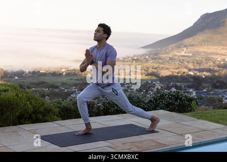 Homme afro-américain pratiquant le yoga fente sur la terrasse de la piscine surplombant la vallée, smartwatch Banque D'Images