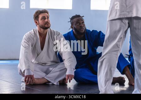 Divers camarades de classe masculins regardant instructeur, assis sur des tapis de dojo portant un sig blanc et bleu Banque D'Images
