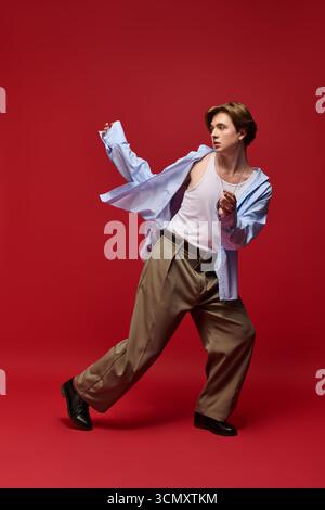 Un jeune homme élégant modèle sa chemise bleue et son pantalon ample sur un fond rouge éclatant. Banque D'Images