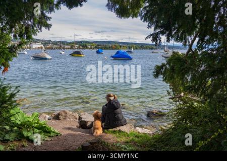Vue naturelle de la fenêtre sur le lac de Zurich (Zurichsee) et femme avec son chien assis sur un rivage à Zurich, Suisse. Banque D'Images