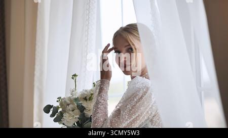 Élégante mariée blonde serrant le bouquet de mariage blanc, rayonnant le bonheur pendant les préparations de jour de mariage Banque D'Images