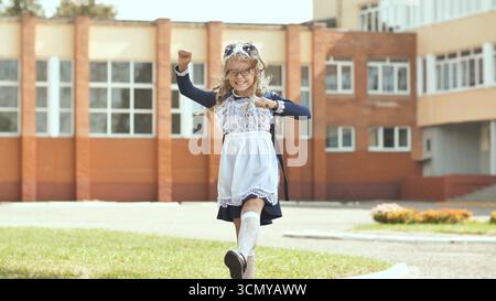 Heureuse écolière dansant devant le bâtiment de l'école, profitant de son éducation et de sa réussite scolaire Banque D'Images