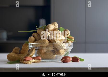 Détail d'un bol en verre avec un tas d'arachides décortiquées sur un banc de cuisine blanc avec des arachides ouvertes montrant les graines dans une cuisine sombre. Vue de face. Banque D'Images