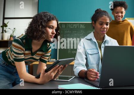 Groupe multiethnique d'adolescents collaborant sur un projet numérique, fille noire utilisant un ordinateur portable tandis que fille caucasienne tenant une tablette, un autre adolescent noir debout derrière eux souriant Banque D'Images