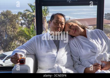 Couple de personnes âgées diversifié assis sur le banc près de la fenêtre dans le spa portant des peignoirs blancs tenant des boissons rouges. Romance, détente, luxe, sérénité, confort, co Banque D'Images