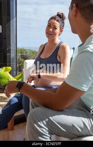 Couple assis sur le balcon donnant sur l'océan, portant des vêtements de sport et smartwatches avec rouleau en mousse. Grossesse, relaxation, bien-être, plein air, style de vie Banque D'Images