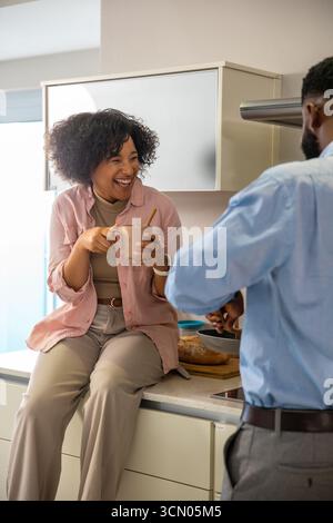 Couple diversifié riant et bavardant dans la cuisine à la maison avec tasse en céramique, pain et poêle à frire. Confortable, domestique, candide, candidshot, faire la maison, kitc Banque D'Images
